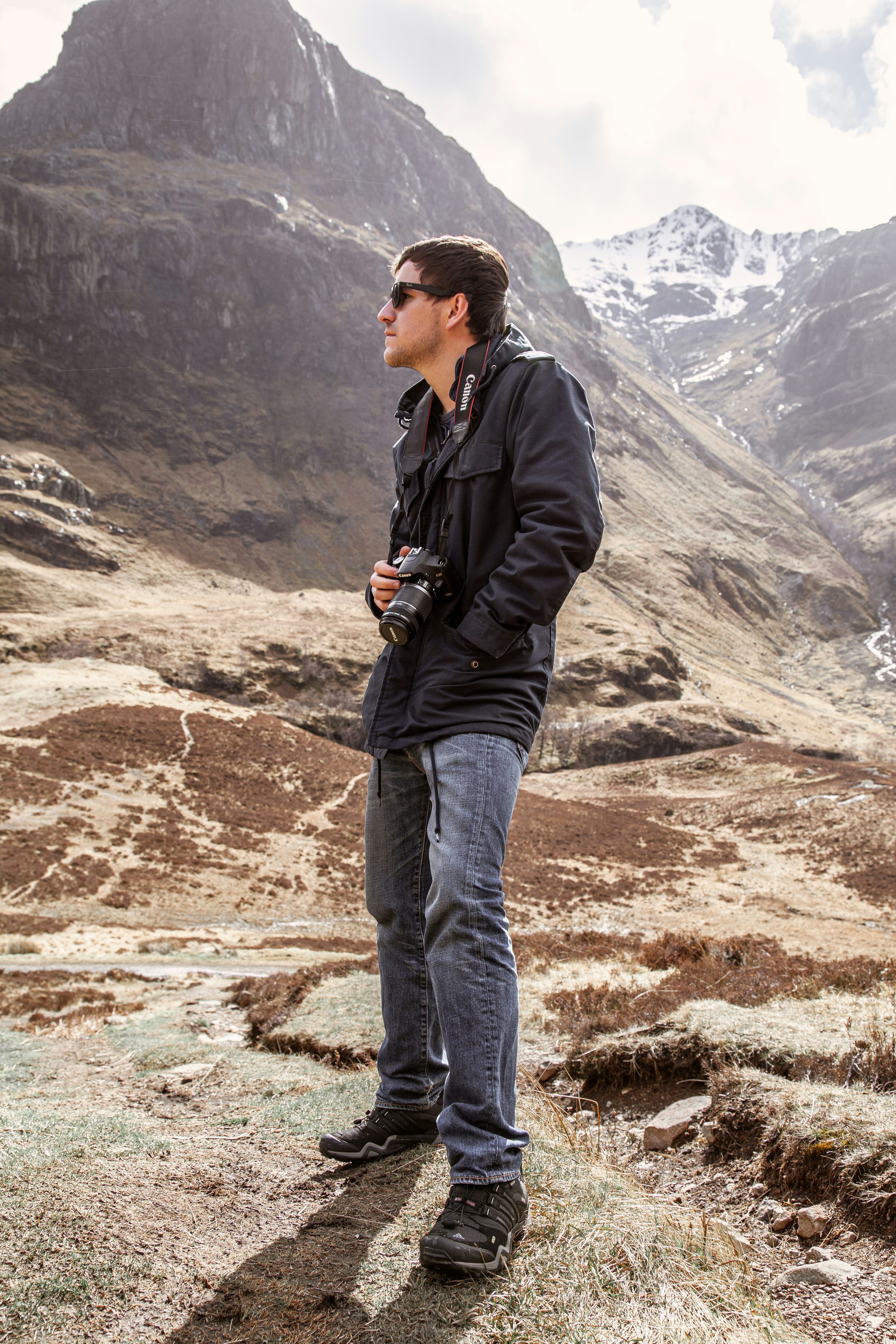 man in black jacket and blue denim jeans holding black dslr camera standing on brown rock